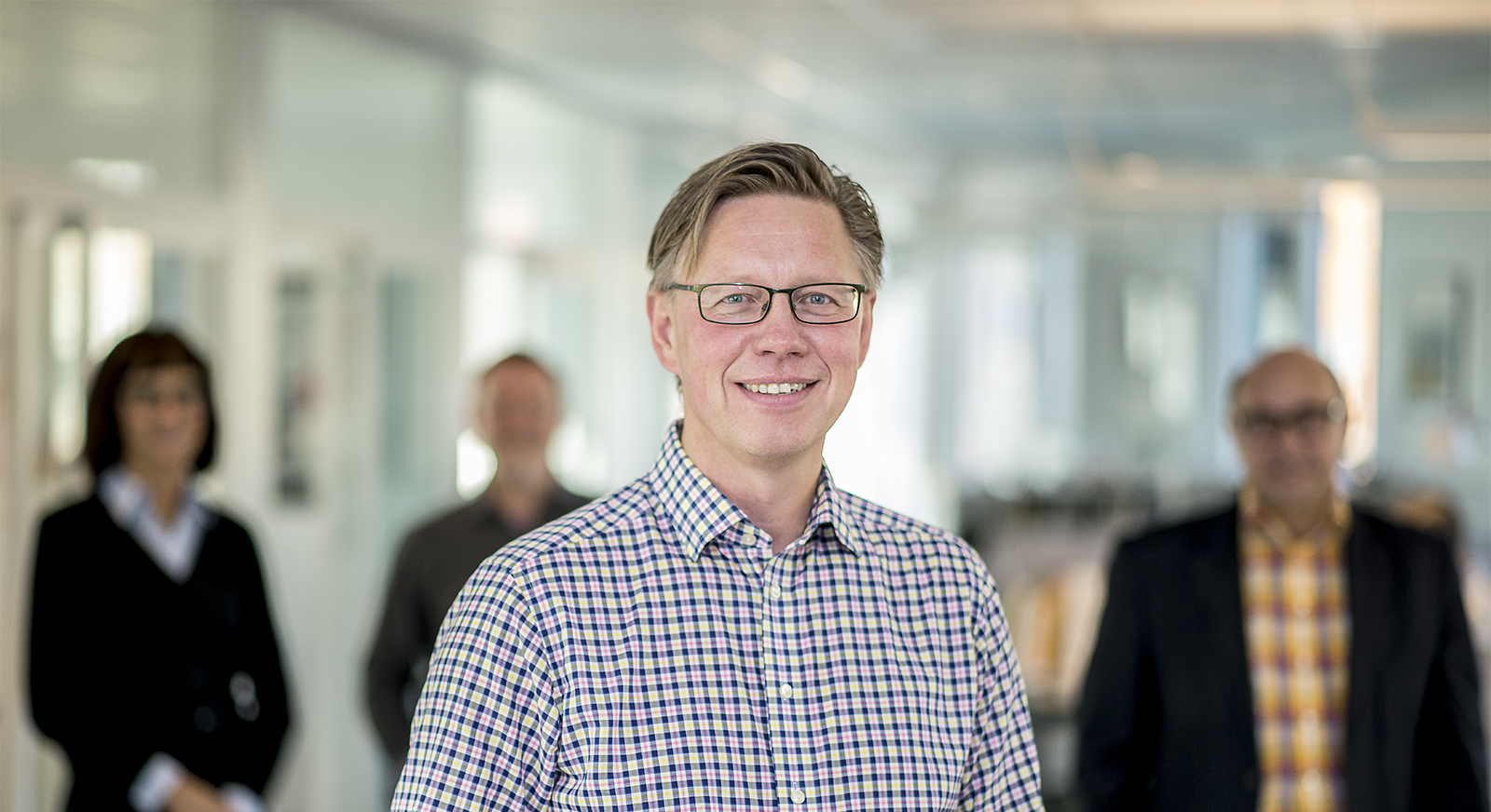 Man med glasögon står leendes mot kameran. I bakgrunden syns 3 suddiga personer.